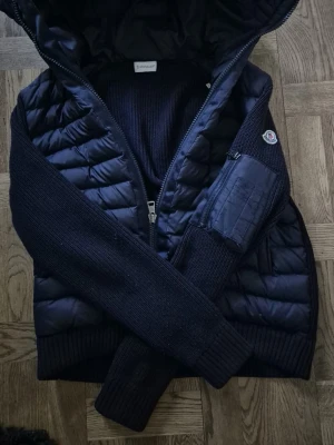 Mörkblå cardiganjacka med huva - Snygg mörkblå cardiganjacka från Moncler med stickade ärmar och quiltad front. Jackan har en praktisk huva, dragkedja framtill och en ficka med dragkedja på ärmen. Perfekt för lager-på-lager och streetwear-vibe.