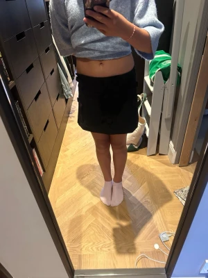 Svart minikjol med rosetter - Säljer en svart minikjol med fem dekorativa rosetter framtill. Kjolen har en rak, något A-linjeformad passform och är sydd i ett slätt tyg. Perfekt för dig som gillar söta detaljer och vill ha en trendig look.