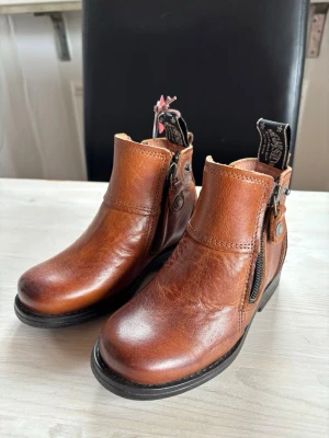 Bruna läderboots - Sneaky Steve - Helt nya snygga bruna boots i äkta läder från Sneaky Steve med dragkedja på sidan och dekorativa nitar. Skorna har en låg klack och rund tå, samt en robust sula i svart. Perfekta för dig som gillar klassisk stil med en modern twist.