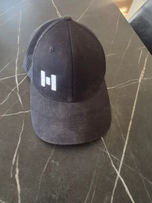 Svart meshkeps med vit logga - Säljer en svart keps från Flexfit med mesh på sidorna och bak, samt en vit broderad logga framtill. Kepsen har böjd skärm och justerbar passform. Perfekt för en avslappnad streetwear-look.