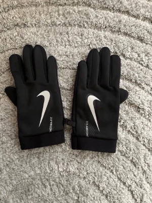 Svarta Nike Therma-FIT handskar - Snygga svarta handskar från Nike med Therma-FIT-teknologi. Handskarna har en vit Nike-logga på ovansidan och är tillverkade i ett mjukt och stretchigt material som håller händerna varma. Perfekta för kyliga dagar och sportiga outfits.