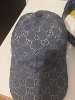 Blå GG-mönstrad keps från Gucci - Snygg blå keps från Gucci med det klassiska GG-monogrammet över hela kepsen. Justerbar rem baktill i matchande färg och böjd skärm. Materialet ser ut att vara tåligt tyg med exklusiv känsla. Perfekt för dig som vill ha en trendig accessoar.