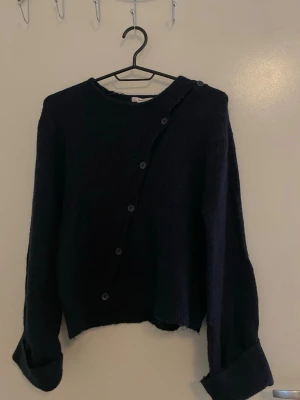 Mörkblå stickad tröja Gina Tricot - Snygg mörkblå stickad tröja från Gina Tricot med asymmetrisk knäppning framtill. Tröjan har långa ärmar och en lite lösare passform, perfekt för lager på lager. Materialet känns mjukt och mysigt, och knapparna ger en unik touch.