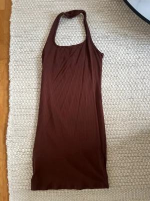 Brun ribbad halterneckklänning - Säljer en brun ribbad klänning med halterneck och tight passform. Klänningen är ärmlös och har en kort längd, perfekt för varma dagar. Materialet är mjukt och stretchigt, vilket gör den bekväm att bära.