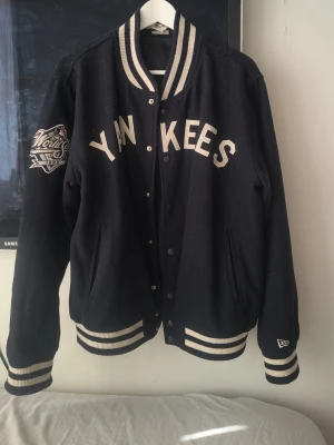 Svart Yankees bomberjacka - Snygg svart bomberjacka med vita detaljer och Yankees-tryck fram och stort NY-logga bak. Jackan har ribbade muddar och krage i svartvitt, samt broderad patch på ärmen. Perfekt för dig som gillar sportig streetstyle.