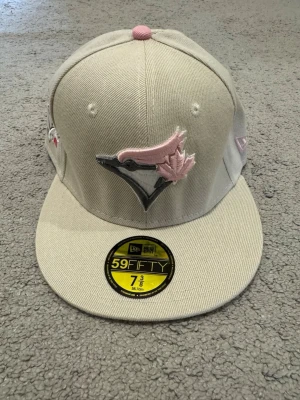Beige New Era 59FIFTY Toronto Blue Jays keps - Snygg beige 59FIFTY keps från New Era med Toronto Blue Jays logga i rosa och grått framtill. Platt skärm och broderade detaljer på sidan med rosa inslag. Klassisk snapback-stil i bomullsmaterial, perfekt för dig som gillar streetwear och sportig look.