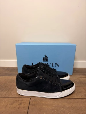 Lanvin cap toe  - Snygga svarta sneakers med ovandel i mocka och glansiga lackdetaljer på tå och häl. Skorna har svarta skosnören och vit sula som ger en clean kontrast. Perfekta för dig som gillar en stilren men ändå edgy look. Storlek uk 6/ 41 men passar även 42. Obs endast skorna ingår 