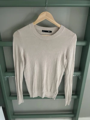 Beige stickad tröja från 157 - Snygg beige stickad tröja från 157 med rund halsringning och långa ärmar. Tröjan har en enkel och stilren design med ribbade muddar vid hals, ärmslut och nederkant. Perfekt att matcha med jeans eller kjol för en clean look.