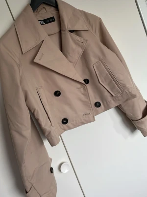 Kort beige trenchcoat från ZARA - Snygg kort trenchcoat från ZARA i beige färg. Jackan har dubbelknäppning med svarta knappar, bred krage och två framfickor. Trendig croppad modell som passar perfekt till jeans eller kjol. Tillverkad i ett slätt tyg som ger en stilren look.
