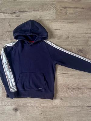Marinblå hoodie från GANT med ränder - Snygg marinblå hoodie från GANT med vita och röda ränder längs ärmarna och GANT-logga på banden. Klassisk huva och magficka framtill. Tillverkad i mjuk bomull, perfekt för en avslappnad och sportig stil.