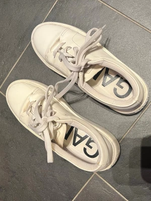 Vita sneakers från Ganni - Säljer ett par stilrena vita sneakers från ganni med tjock sula. Skorna har vita snören och är tillverkade i slätt material som ger en clean look. I toppen skick, bara lite smutsiga på undersidan men det syns ju inte