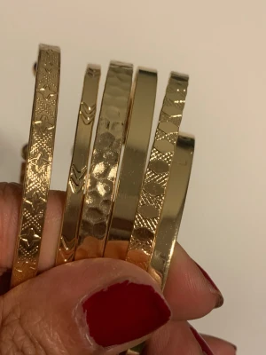Guldiga armband med mönster - Säljer ett set med sex guldiga armband i metall. Armbanden har olika ingraverade mönster som hjärtan, prickar och geometriska former. De är stela och öppna i ändarna vilket gör dem lätta att ta på och av. Perfekta för att ge din outfit en extra touch av stil.