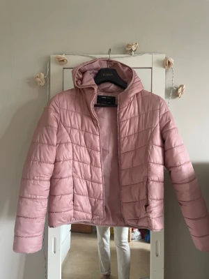 Ljusrosa pufferjacka med huva - Säljer en ljusrosa pufferjacka med huva och dragkedja framtill. Jackan har quiltad design och två sidofickor. Perfekt för kalla dagar och ger en snygg, avslappnad look. Materialet är syntet och jackan är lätt vadderad för extra värme.