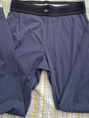 Mörkblå leggings från Alo - Snygga mörkblå leggings från Alo med bred svart midjeresår och diskret logga framtill. Tillverkade i ett stretchigt och mjukt material som sitter skönt på kroppen. Perfekta för träning eller chill.