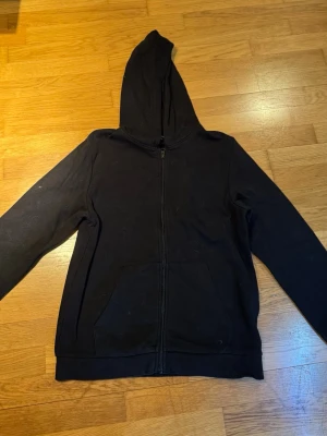 Svart hoodie med dragkedja från Take Off - Klassisk svart hoodie från Take Off med dragkedja framtill och två stora fickor. Hoodien har en rymlig huva och är tillverkad i mjukt bomullsmaterial. Perfekt för chill dagar eller när du vill ha en enkel och stilren look.