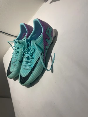 Nike turkosa och lila fotbollsskor - Säljer ett par Nike fotbollsskor i en fräsch turkos färg med lila och vita detaljer samt svarta Nike-loggor. Skorna har snörning och dobbar för bra grepp på planen. Perfekta för dig som vill sticka ut på matchen!