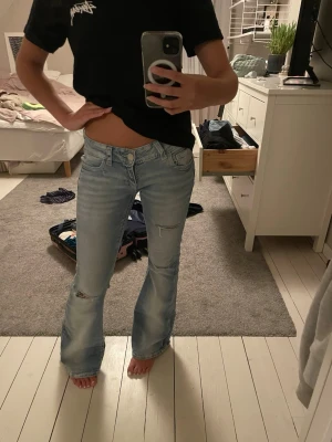 Ljusblå bootcut jeans med slitningar - Säljer ett par ljusblå bootcut jeans med slitningar på knäet och detaljerade bakfickor med knappar. Jeansen har låg midja och klassisk femficksdesign. Perfekta för en avslappnad och trendig look.