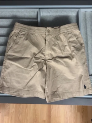 Beige shorts från Ralph Lauren - Klassiska beige shorts från Ralph Lauren med elastisk midja och knappstängning. Shortsen har sidofickor och en liten broderad logga i mörkblått på benet. Perfekta för varma dagar och enkel att matcha med olika stilar.helt nya