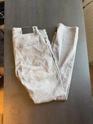 Ljusgrå raka jeans - Säljer ett par ljusgrå jeans med rak passform och klassisk femficksdesign. Jeansen har en enkel look utan slitningar eller dekorationer och är tillverkade i mjuk bomull. Perfekta för dig som gillar en clean och stilren stil.