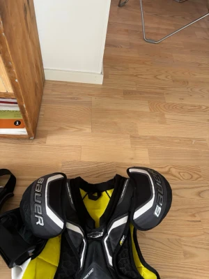 Svart och gul hockeyaxelskydd Bauer - Säljer ett par hockeyaxelskydd från Bauer i svart, vitt och gult. Skydden har vadderade axelpartier, meshpaneler för ventilation och justerbara remmar för bästa passform. Perfekt för dig som vill ha bra skydd på isen.