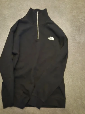 Svart halfzip från The North Face - Svart långärmad tröja från The North Face med halv dragkedja och hög krage. Klassisk logga på bröstet i vitt. Perfekt för lager-på-lager och chill stil. Materialet ser ut att vara mjukt och bekvämt, troligen bomull eller bomullsblandning. 