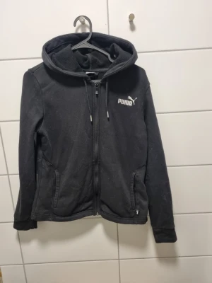 Svart hoodie från Puma med dragkedja - Snygg svart hoodie från Puma med dragkedja framtill och två fickor. Klassisk logga på bröstet och justerbar huva med snören. Tillverkad i mjukt material som känns skönt mot huden. Perfekt för en avslappnad och sportig stil.