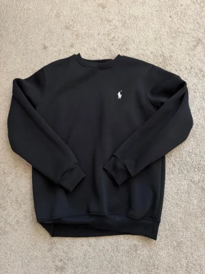 Svart sweatshirt från Polo Ralph Lauren - Klassisk svart sweatshirt från Polo Ralph Lauren med rund halsringning och vit broderad logga på bröstet. Tröjan har ribbade muddar vid ärmslut och nederkant, perfekt för en clean och stilren look.