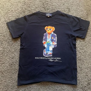 Ralph Lauren  t-shirt  - Snygg marinblå t-shirt från Ralph Lauren med ikoniskt Polo Bear-tryck framtill. T-shirten har rund hals och korta ärmar, perfekt för en avslappnad och trendig look. Tillverkad i mjuk bomull för skön komfort. Priset kan diskuteras 