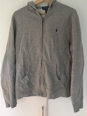 Grå zip up hoodie från Polo Ralph Lauren - Klassisk grå zip up hoodie med dragkedja från Polo Ralph Lauren. Tröjan har huva, två fickor framtill och den ikoniska lilla loggan broderad på bröstet. Tillverkad i mjukt bomullsmaterial och har ribbade muddar vid ärmslut och nederkant.