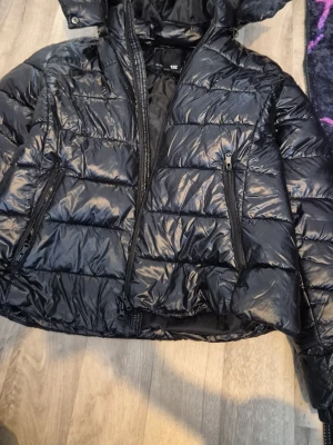 Black Puffer Jacket - This is a black puffer jacket from the brand 157 (established 1999), size XL. The jacket features a shiny, quilted design with a hood and zippered pockets on both sides. It appears to be in good condition and is suitable for cold weather. Jag är svensk*