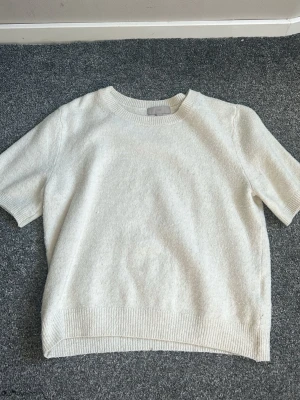 Vit stickad t-shirt med kort ärm - En stilren vit stickad t-shirt med korta ärmar och rund hals. Tröjan har ribbade detaljer vid hals, ärmslut och nederkant. Perfekt för en clean och enkel look. Materialet ser mjukt och bekvämt ut.