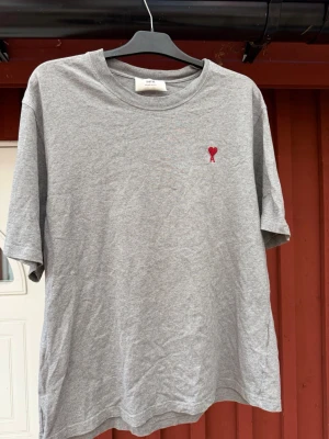 Grå t-shirt från Ami Paris - Snygg grå t-shirt från Ami Paris med röd broderad logga på bröstet. Klassisk rund hals och avslappnad passform. Perfekt till jeans eller shorts för en clean och enkel look.