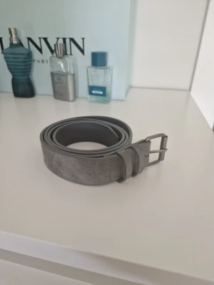 Grått mockabälte  - Stilrent grått bälte i mocka med klassiskt metallspänne. Perfekt accessoar för att lyfta din outfit med en enkel men snygg detalj. Bältet har en slät yta och passar till både jeans och byxor. 