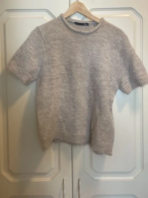 Beige stickad t-shirt - Mysig beige stickad t-shirt med korta ärmar och rund hals. Tröjan har en mjuk och fluffig struktur samt ribbstickade muddar vid ärmslut och nederkant. Perfekt för lager på lager och enkel att matcha med olika stilar.