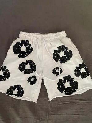 Vita shorts med svart mönster - Unika vita shorts med elastisk midja och snörning. Shortsen har stora svarta blommiga tryck över hela plagget och råa kanter vid bensluten. Perfekta för dig som vill sticka ut med en cool och avslappnad stil.