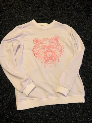 Vit stickad Kenzo tröja med tiger - Säljer en vit stickad tröja från Kenzo med ikoniskt rosa tigerbroderi och texten 'KENZO PARIS' på bröstet. Tröjan har rund halsringning, långa ärmar och ribbade muddar. Perfekt för dig som vill ha en trendig och unik look.