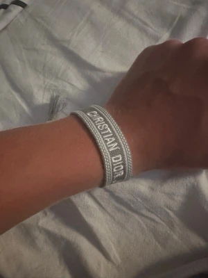 Grått Christian Dior tygarmband - Snyggt grått armband från Christian Dior i vävt tyg med broderad logga. Armbandet har justerbar knytning med tofsar i ändarna för en cool och trendig look. Perfekt accessoar för att lyfta din outfit.
