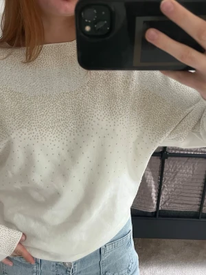 Vit stickad tröja med strassdetaljer - Snygg vit stickad tröja med glittrande strassdetaljer över bröstet, axlarna och ärmarna. Tröjan har en avslappnad passform, rund halsringning och långa ärmar. Perfekt för dig som vill ha något extra till din outfit.