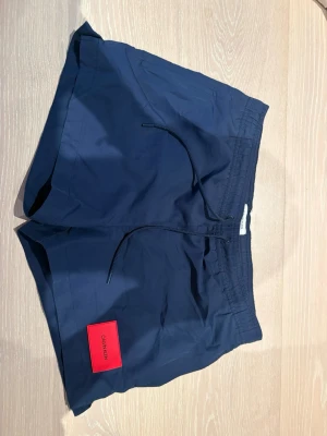 Mörkblå badbyxor med röd detalj - Snygga mörkblå badbyxor med elastisk midja och dragsko. På vänster ben finns en röd patch med texten CALVIN KLEIN. Badbyxorna har sidofickor och en bakficka med liten etikett. Perfekta för stranden eller poolen. (Pris går att diskutera)