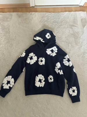 Mörkblå denim tears hoodie - Snygg mörkblå hoodie från denim tears med stora vita blommönster över hela tröjan. Hoodien har en klassisk känguruficka framtill och en rymlig huva. Perfekt för dig som gillar en unik och trendig stil. Materialet känns mjukt och bekvämt, troligen bomull eller bomullsblandning. Strlk S men passar bättre som M