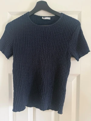 Mörkblå ribbad t-shirt från Zara - En mörkblå ribbad t-shirt från Zara med korta ärmar och rund hals. Materialet har en strukturerad, stretchig känsla som ger en unik look. Perfekt till jeans eller kjol för en enkel men snygg outfit.