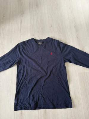 Mörkblå långärmad t-shirt Polo Ralph Lauren - Snygg mörkblå långärmad t-shirt från Polo Ralph Lauren med klassisk röd logga broderad på bröstet. Tillverkad i mjuk bomull och har en normal passform. Perfekt för en clean och stilren look.