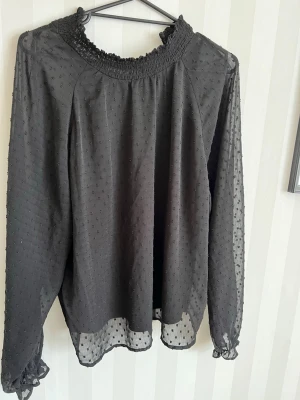 Svart  blus med prickar - Snygg svart blus i tunt material med små prickar. Blusen har långärmad design, smockad halsringning och volangdetaljer vid ärmslut. Perfekt för dig som gillar trendiga och feminina plagg. Blusen är täckande framtill men genomskinlig i ärmarna och i ryggen🤍Köptes för några år sedan, men har mest använt vid speciella tillfällen. Är i gott skick🤍Från JDY, i storlek L, men passar även M!