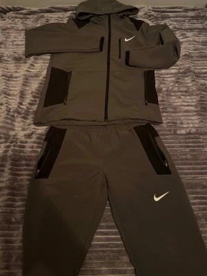 Nike grått tracksuit set - Säljer ett grått loungewear set från Nike med svartvita detaljer. Setet består av en hoodie med dragkedja, fickor och Nike-logga samt matchande byxor med resår i midjan och dragkedjeförsedda fickor. Perfekt för chill dagar hemma eller på språng. Smutsig på bröstet.