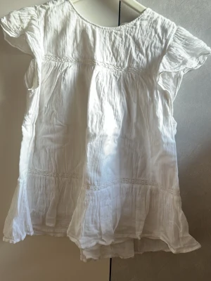 Vit blus med volang och kort ärm - Luftig vit blus i bomull med volangdetaljer och korta ärmar. Blusen har en lätt rynkad struktur och v-ringning framtill. Perfekt för en avslappnad och somrig look. Storlek 170.