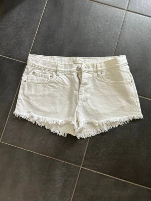 Vita jeansshorts med fransig kant - Säljer ett par vita jeansshorts med fransig kant och klassisk femficksdesign. Shortsen har bälteshällor, knapp- och dragkedjestängning samt råa kanter nedtill för en avslappnad look. Perfekta för sommaren!