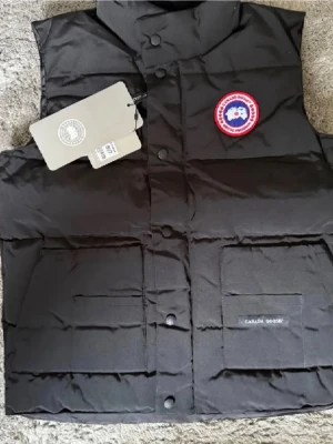 Svart dunväst från Canada Goose - Snygg svart dunväst från Canada Goose med klassisk logotyp på bröstet och två stora fickor framtill. Västen har hög krage och stängs med tryckknappar. Perfekt för lager-på-lager och håller dig varm under kyliga dagar.