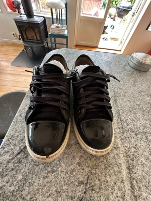 Svarta sneakers med lackdetaljer - Ett par snygga svarta lanvin Captoe tjej modell som är i bra skick med allt kvar passar till alla kläder och är bra till hösten men har dock ett litet håll i captoen på högra skon men gör inte så mycket 