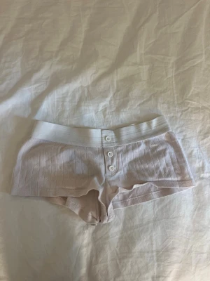 Ljusrosa ribbade pyjamasshorts - Säljer ett par ljusrosa pyjamasshorts med ribbad struktur och bred resår i midjan. Shortsen har dekorativa knappar framtill och är tillverkade i ett mjukt, stretchigt material som passar perfekt för mysiga hemmakvällar.