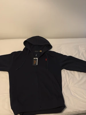 Svart hoodie från Polo Ralph Lauren - Snygg svart hoodie från Polo Ralph Lauren med dragkedja framtill och klassisk röd logga broderad på bröstet. Huva med dragsko och mjukt material som känns skönt mot huden. Perfekt för en avslappnad och stilren look.